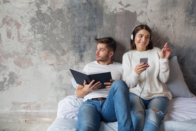 Young smiling couple sitting on bed at home in casual outfit reading book wearing jeans, man reading book, woman listening to music on headphones, spending romantic time together