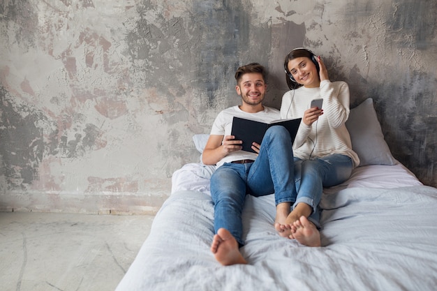 Young smiling couple sitting on bed at home in casual outfit reading book wearing jeans, man reading book, woman listening to music on headphones, spending romantic time together