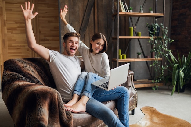 Young smiling couple sitting on couch at home in casual outfit, love and romance, woman and man embracing, wearing jeans, spending relaxing time together, holding laptop, happy emotional