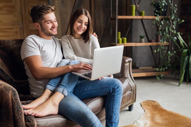 Young smiling couple sitting on couch at home in casual outfit, love and romance, woman and man embracing, wearing jeans, spending relaxing time together, holding laptop