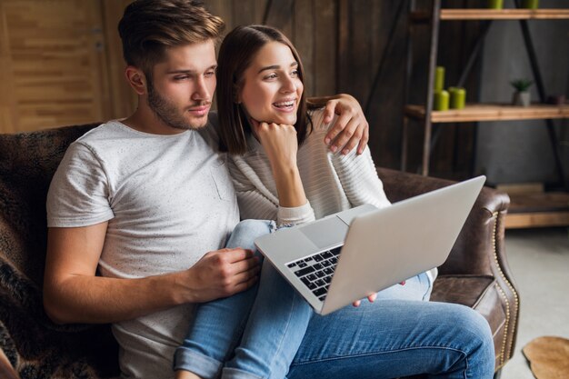 Young smiling couple sitting on couch at home in casual outfit, love and romance, woman and man embracing, wearing jeans, spending relaxing time together, holding laptop