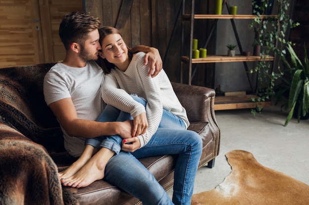 Young smiling couple sitting on couch at home in casual outfit, love and romance, woman and man embracing, wearing jeans, spending relaxing time together