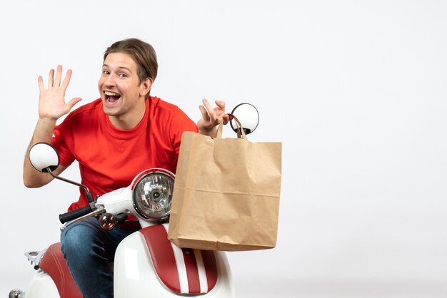 Young smiling courier guy in red uniform sitting on scooter holding paper bag showing five on white wall