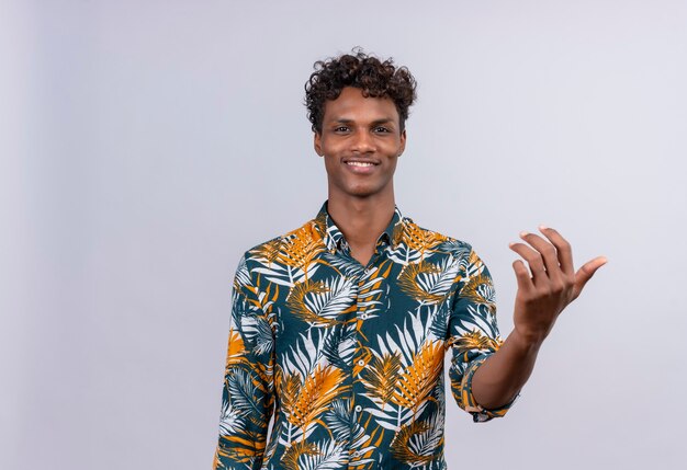 A young smiling dark-skinned man with curly hair in leaves printed shirt  and calling closer with hand gesture 