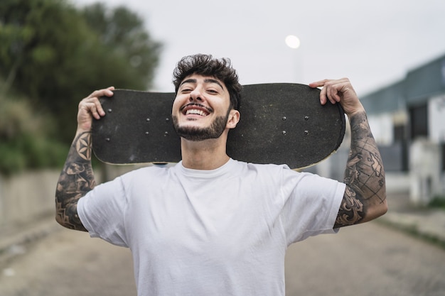 거리의 흐릿한 환경에서 스케이트를 들고 웃고 있는 젊은 유럽 멋진 남성