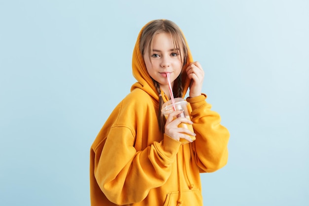 Молодая улыбающаяся девушка в оранжевой толстовке с капюшоном пьет сок из пластиковой чашки, счастливо глядя в камеру на синем фоне