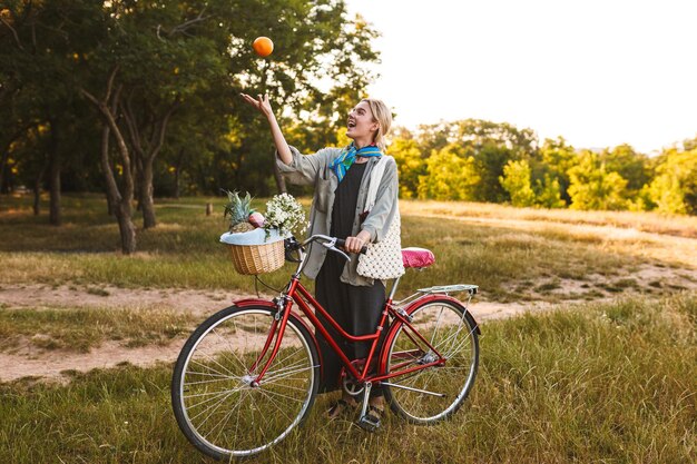 Young smiling girl with red bicycle and wildflowers and fruits in basket happily playing with orange in park