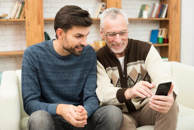 Young smiling guy and aged cheerful man using mobile phone on settee