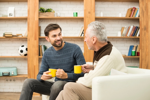 Young smiling guy and aged man with cups on sofa