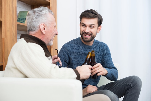 Young smiling guy clanging bottles with aged man on sofa