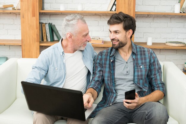 Young smiling guy with smartphone pointing at monitor of laptop on legs of aged man on sofa