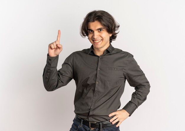 Молодой улыбающийся красивый кавказский мужчина указывает вверх и кладет руку на талию, изолированную на белом фоне с копией пространства