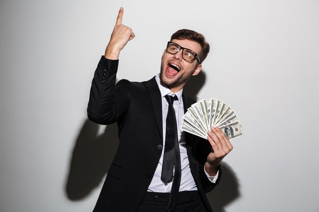 Young smiling handsome man in classic black suit holding bunch of money while pointing with finger upward