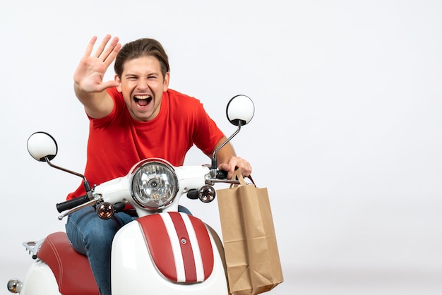 Young smiling happy courier guy in red uniform sitting on scooter holding orders showing five on yellow wall