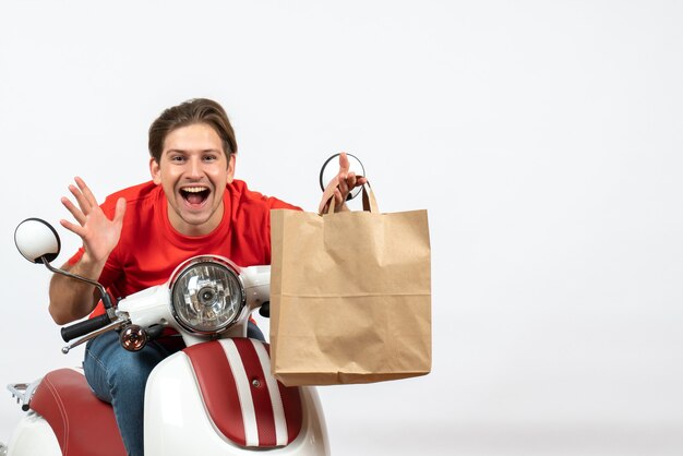 Молодой улыбающийся счастливый курьер в красной форме сидит на скутере и держит бумажный пакет на белой стене