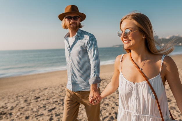Молодой улыбающийся счастливый человек в шляпе и белокурая женщина вместе бегают на пляже на летних каникулах, путешествуя