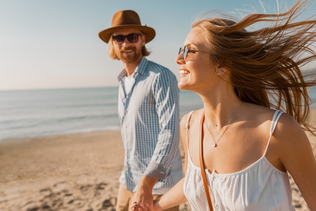 Young smiling happy man in hat and blond woman running together on beach on summer vacation traveling