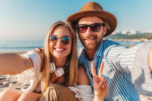 Foto gratuita giovane sorridente felice uomo e donna in occhiali da sole seduti sulla spiaggia di sabbia prendendo selfie foto sulla fotocamera del telefono