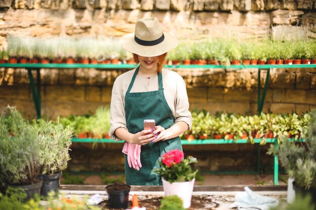 Free photo young smiling lady in apron and hat standing and taking photos of flower in pot on her cellphone while working in greenhouse