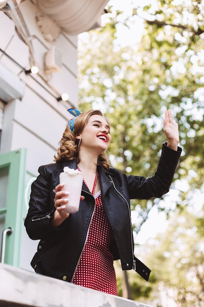 Young smiling lady in leather jacket standing with milkshake in hand and happily looking aside while waving to someone