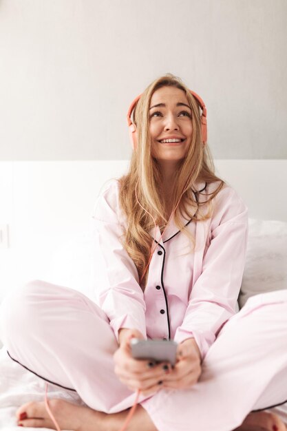 Young smiling lady in sleepwear sitting on bed and listening music in headphones with cellphone in hands at home