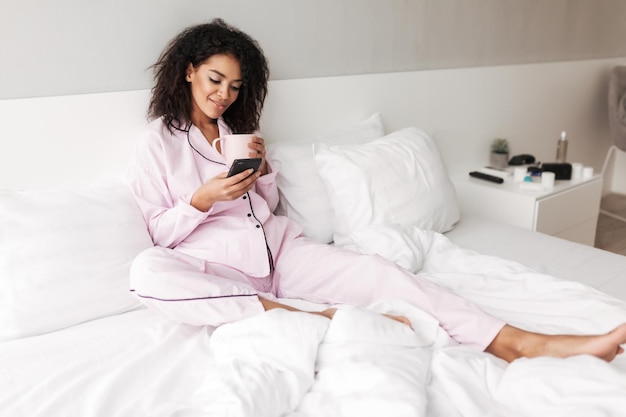 Молодая улыбающаяся дама с темными вьющимися волосами в пижаме сидит дома в постели с чашкой и мобильным телефоном в руках