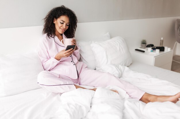 Young smiling lady with dark curly hair in sleepwear sitting in bed with cup and cellphone in hands at home