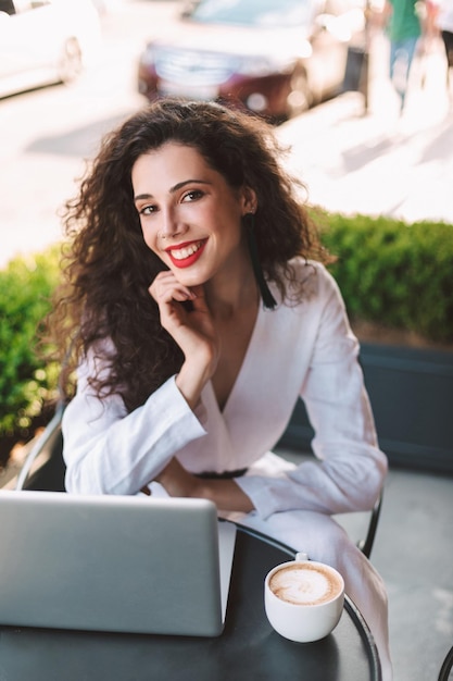 Free photo young smiling lady with dark curly hair in white costume sitting at the table with cup of coffee and laptop while happily looking in camera in cafe on street