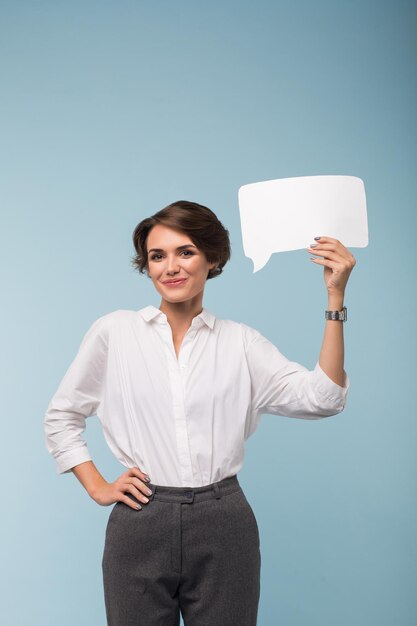 Young smiling lady with dark short hair in shirt and pants joyfully looking in camera holding white paper shape of message in hand near head over blue background isolated