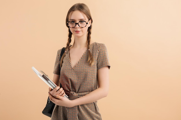 Young smiling lady with two braids in tweed jumpsuit and eyeglasses with black backpack on shoulder holding notepads in hands while dreamily looking in camera over beige background