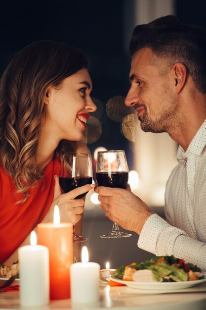 Молодые улыбающиеся любовники смотрят друг на друга и ужинают с вином и едой