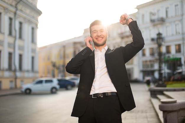 Молодой улыбающийся мужчина в черной куртке и белой рубашке радостно поднимает руку во время разговора по мобильному телефону с прекрасным видом на город на заднем плане