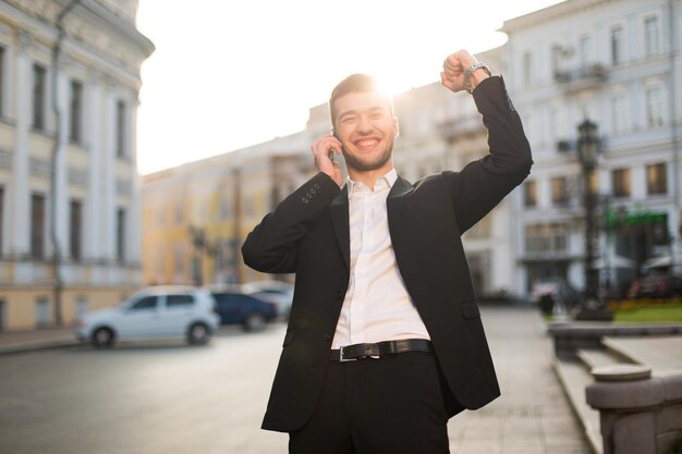 검은색 재킷과 흰색 셔츠를 입은 웃고 있는 젊은 남자는 배경에서 아름다운 도시 전망을 배경으로 휴대폰으로 이야기하면서 행복하게 손을 들어 올립니다