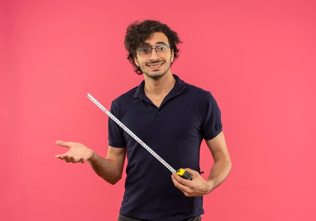 Young smiling man in black shirt with optical glasses holds tape measure isolated on pink wall