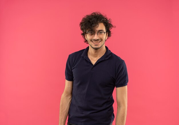 Young smiling man in black shirt with optical glasses looks isolated on pink wall