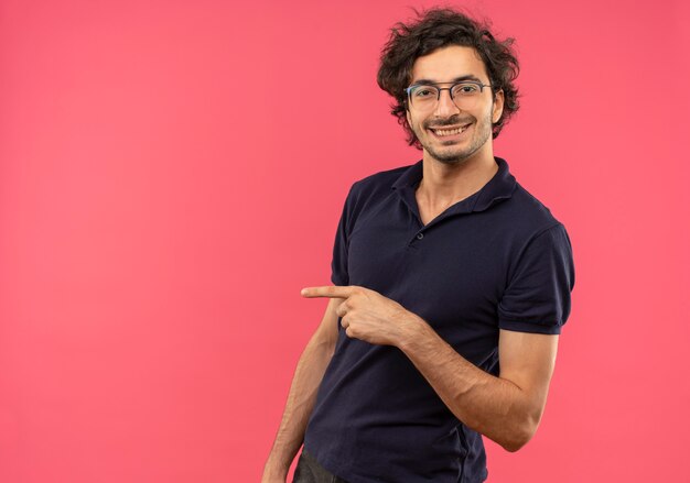 Young smiling man in black shirt with optical glasses points at side isolated on pink wall