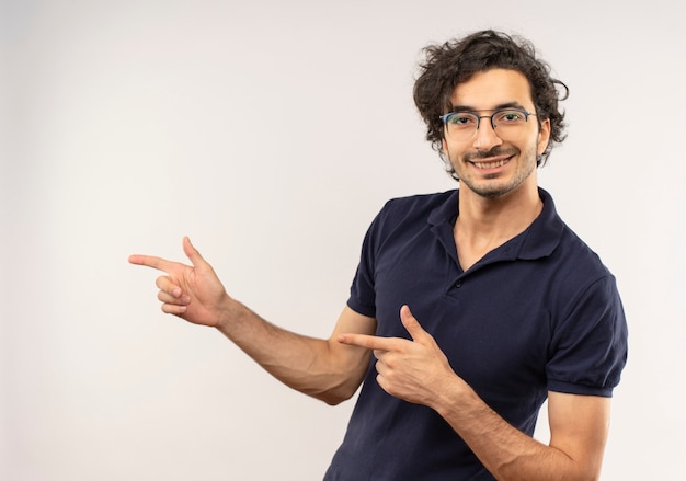 Young smiling man in black shirt with optical glasses points at side and looks isolated on white wall