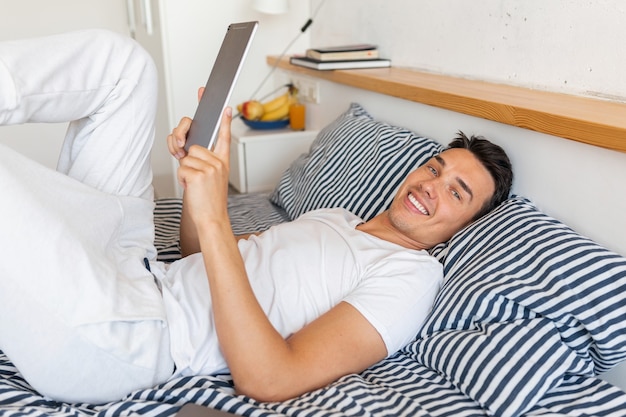 Young smiling man in casual pajamas outfit sitting in bed in morning holding tablet