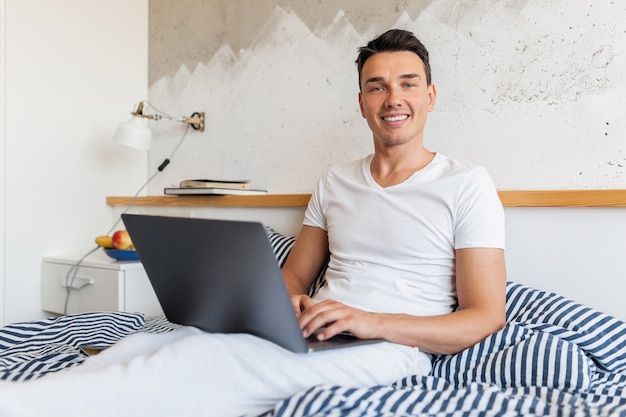 Молодой улыбающийся человек в повседневной пижамной одежде сидит в постели утром, работая на ноутбуке
