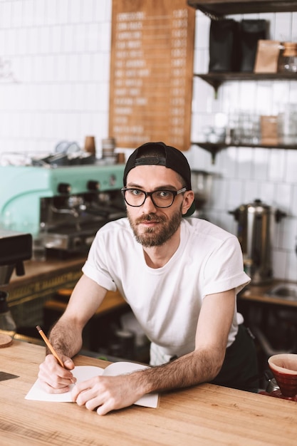 鉛筆とメモ帳でバーカウンターの後ろに立って、カフェで働いている間、夢のようにカメラで見ている眼鏡とキャップの若い笑顔の男