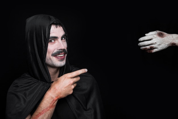 Young smiling man in Halloween costume pointing at corpse hand