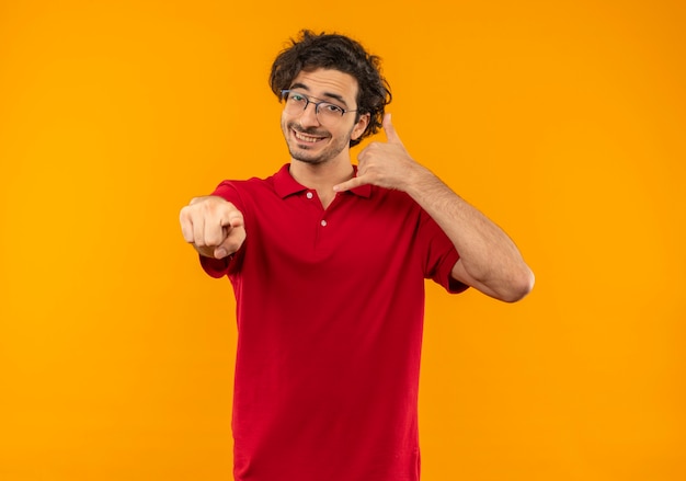 Young smiling man in red shirt with optical glasses gestures call me hand sign and points isolated on orange wall