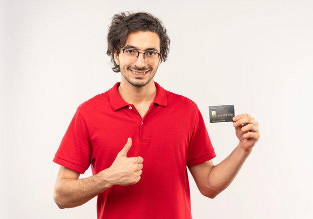 Young smiling man in red shirt with optical glasses holds credit card and thumbs up isolated on white wall