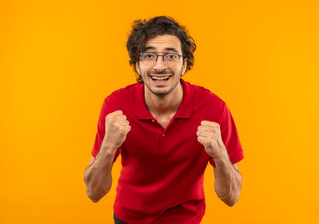 光学メガネと赤いシャツを着た若い笑顔の男は、オレンジ色の壁に隔離された拳を持ち上げます