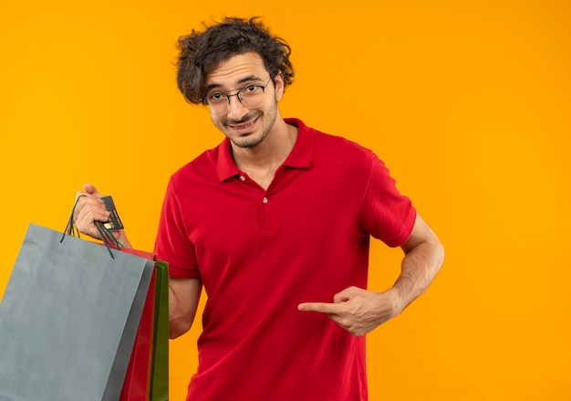 Молодой улыбающийся человек в красной рубашке с оптическими очками держит и указывает на бумажные пакеты, изолированные на оранжевой стене