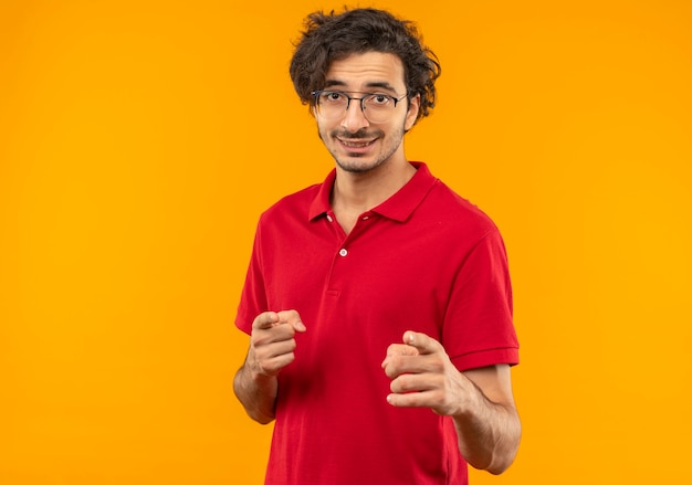 Young smiling man in red shirt with optical glasses points isolated on orange wall