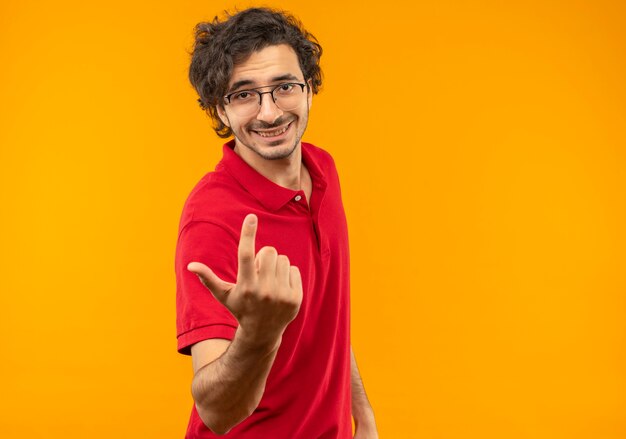 Young smiling man in red shirt with optical glasses points up isolated on orange wall