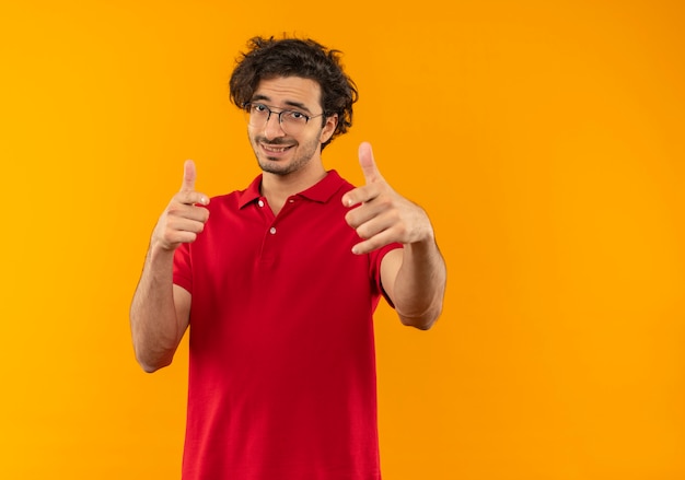 Young smiling man in red shirt with optical glasses points with hands isolated on orange wall