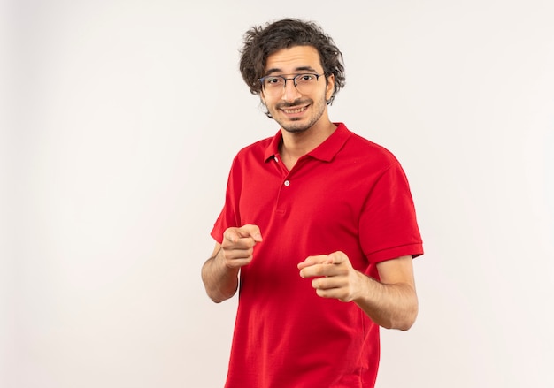 Young smiling man in red shirt with optical glasses points with hands isolated on white wall