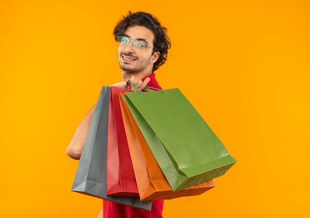 Young smiling man in red shirt with optical glasses stands sideways and holds multicoloured paper bags isolated on orange wall